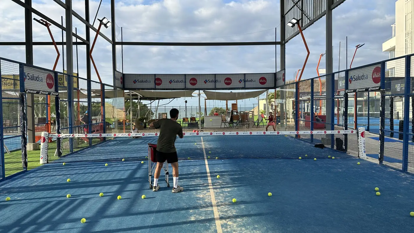 Clase de padel en cancha con el entrenador, canasto de pelotas y el mar al fondo