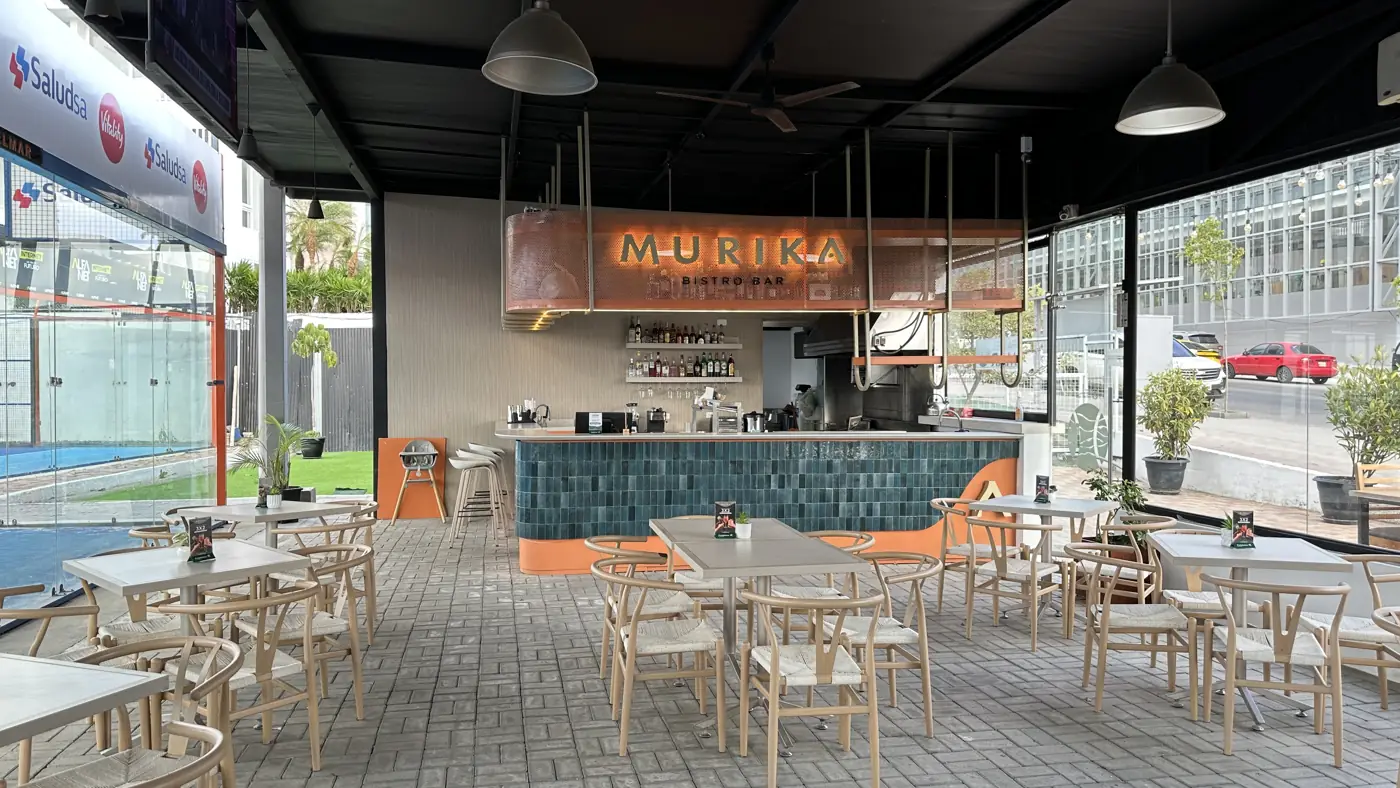 Salón de Murika Bistro Bar con mesas y sillas de madera, la barra al fondo y una cancha de padel a un lado