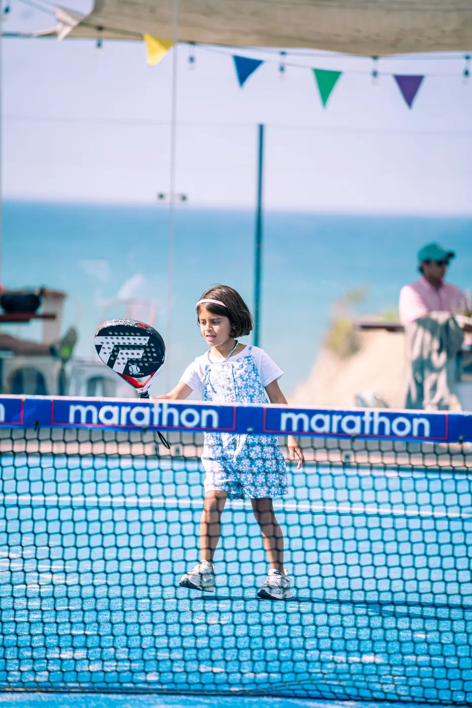 Niña jugando junto a la red de la cancha con su paleta, con el mar al fondo