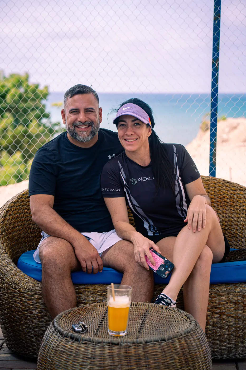 Pareja sonriendo en un sillón de mimbre del jardín, con el mar al fondo y un vaso de cerveza