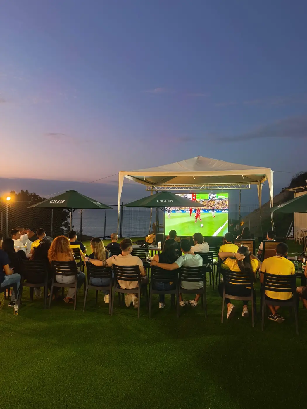 Público viendo un partido al atardecer en Padelmar Gardens, con pantalla grande y el mar al fondo
