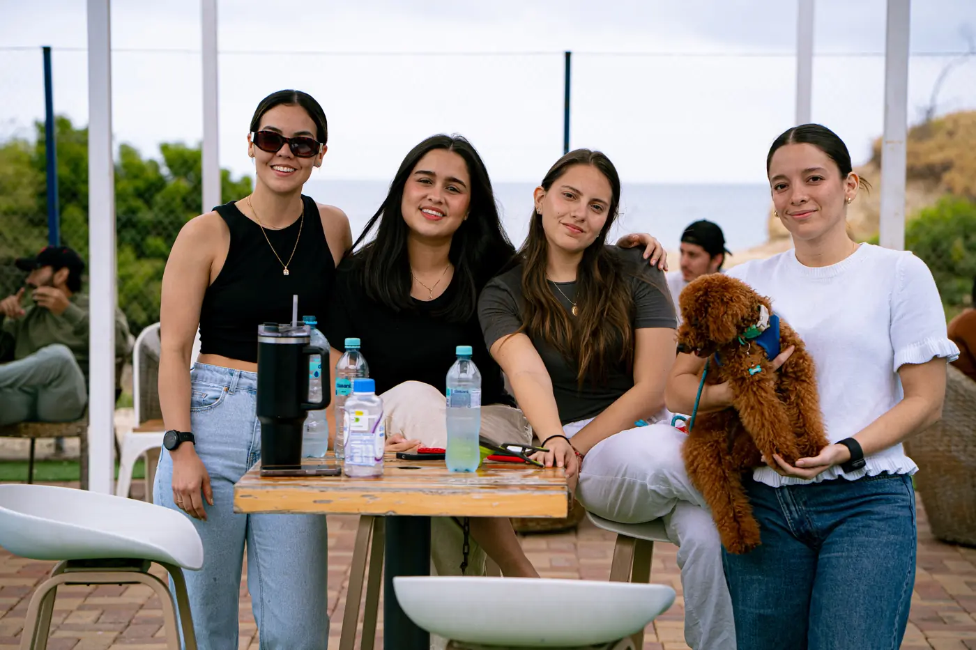 Cuatro amigas sonriendo en una mesa del jardín, una de ellas con un perrito poodle en brazos, con el mar al fondo