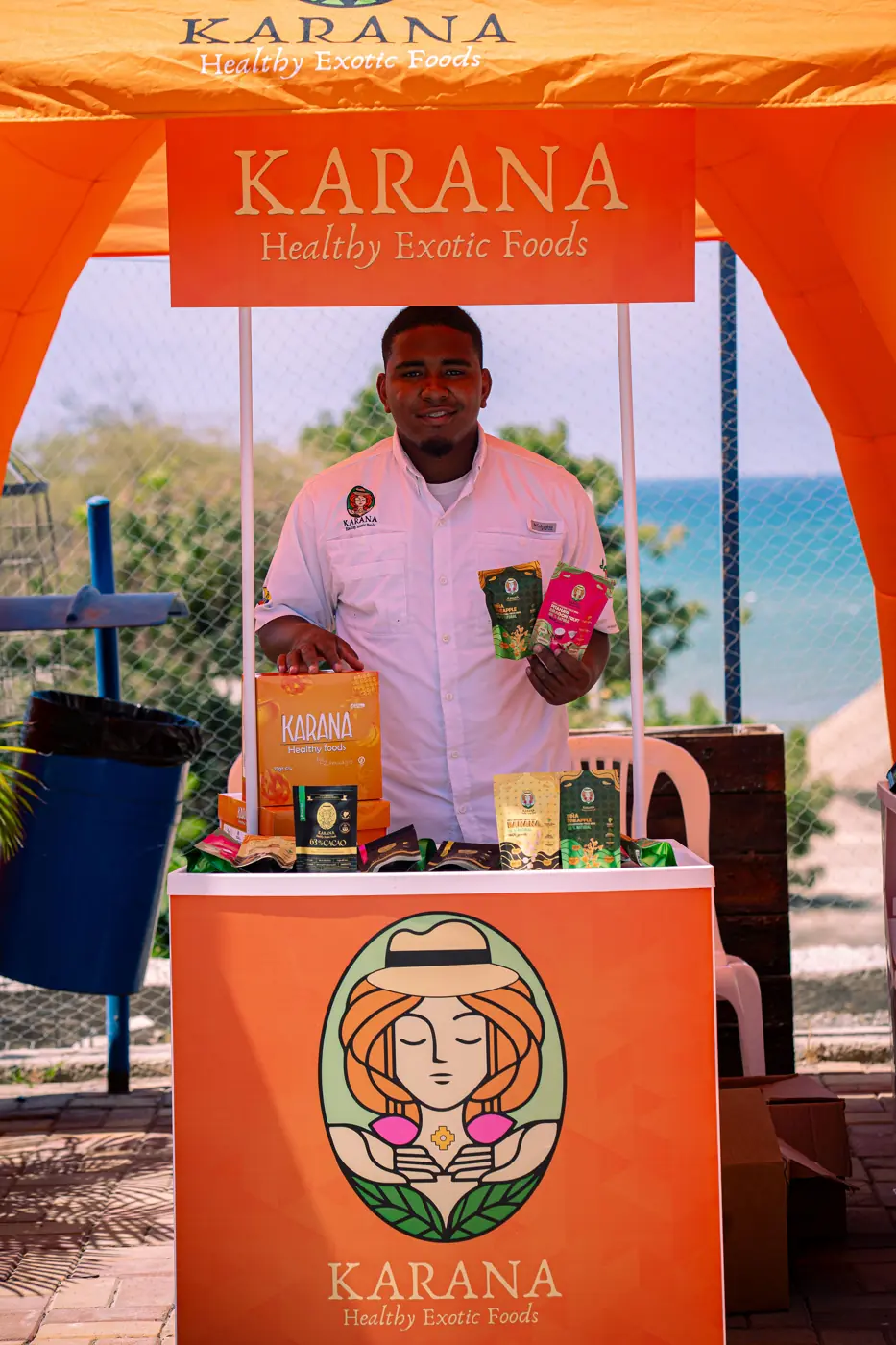 Stand de Karana Healthy Exotic Foods con sus productos y el mar al fondo